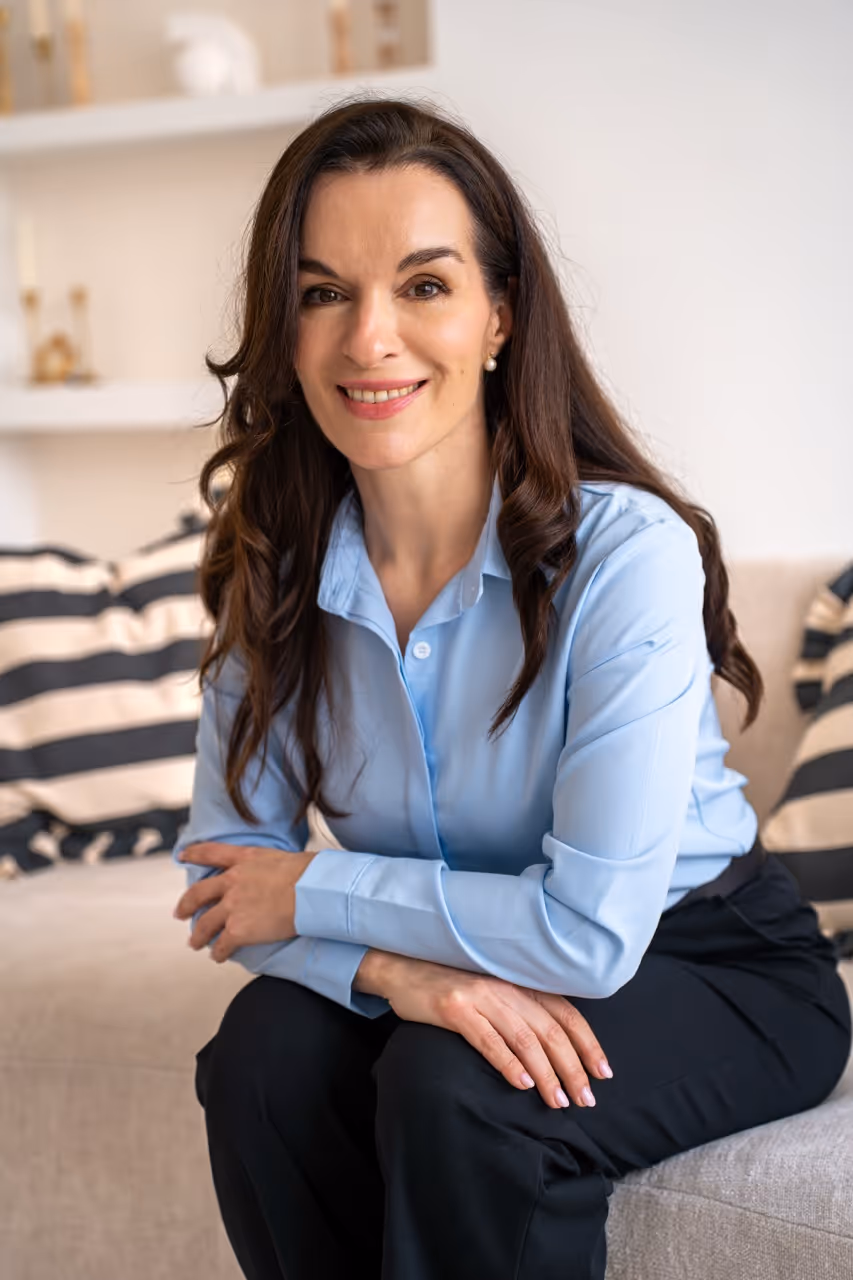 Smiling woman with long dark hair wearing a light blue shirt and black pants sitting on a beige sofa.